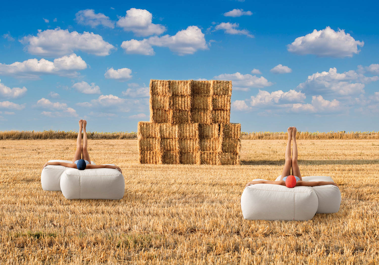 two women in swimsuits sticking legs up in the air on white ottomans in a hay field