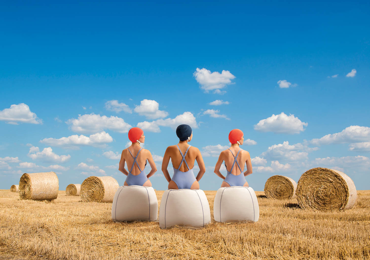 three women in swimsuits sitting on white ottomans in a hay field